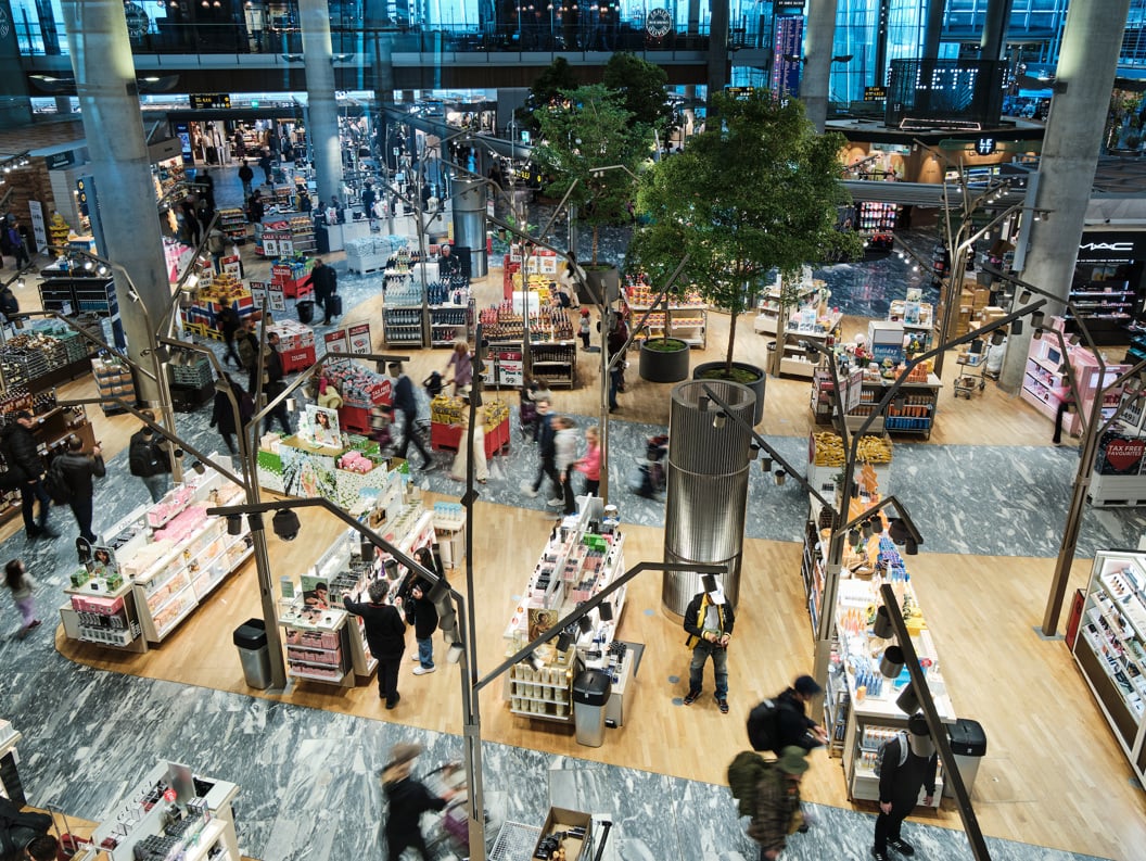 Interiør av en travel flyplassbutikk med folk som handler, omgitt av hylleutstillinger med kosmetikk, parfyme og andre varer. Butikken har moderne innredning med trær og åpen planløsning