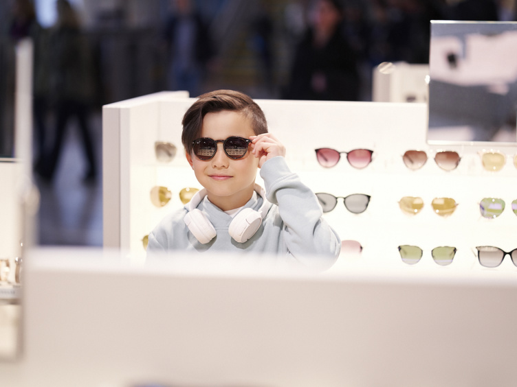 Ung gutt prøver solbriller i butikk, omgitt av utstilte briller, med hodetelefoner rundt halsen.