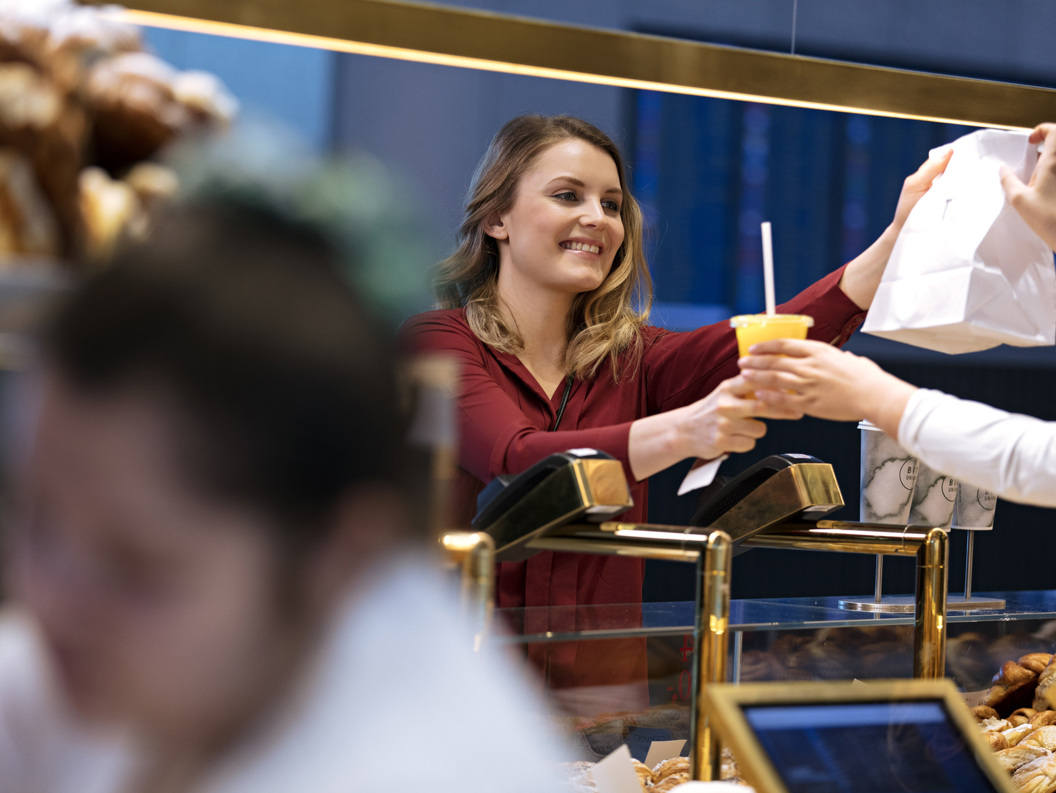 Kvinne i kafé gir kunde en papirpose og en kald drikke over disken.