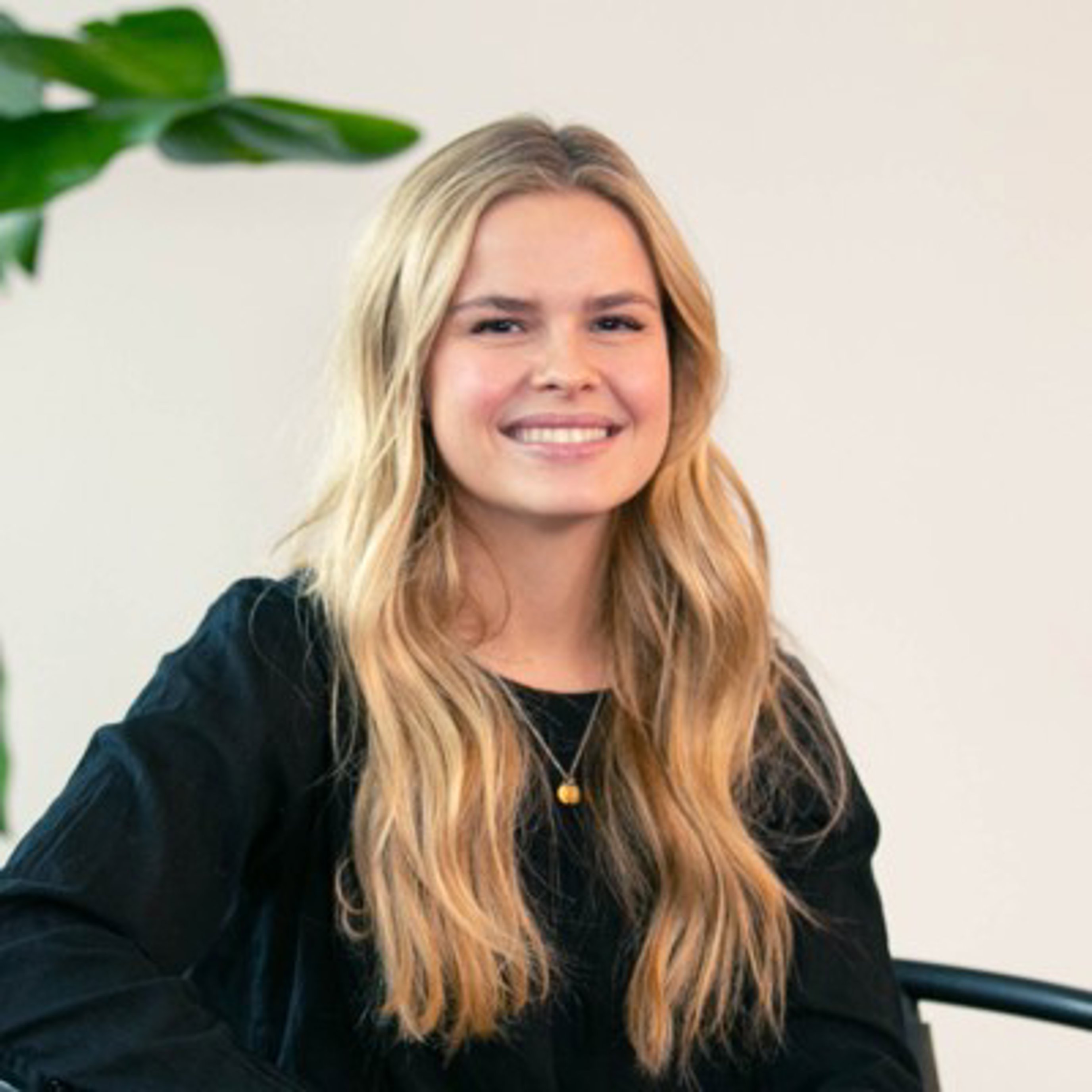 Smilende kvinne med langt blondt hår som har på seg en svart topp, sittende innendørs med en plante i bakgrunnen