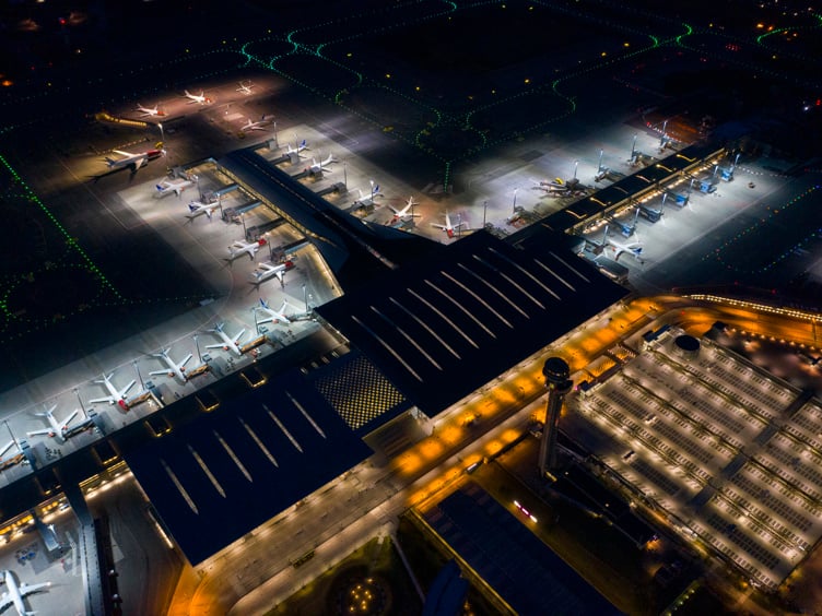 Flyfoto av opplyst flyplass med parkerte fly og terminalbygning om natten.