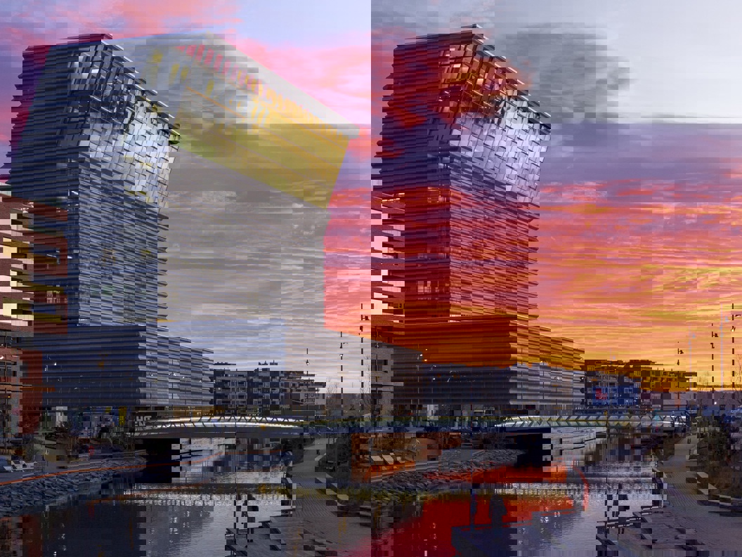 Munchmuseet i Oslo ved solnedgang, med dramatiske rosa og oransje skyer i bakgrunnen, reflektert i vannet nedenfor.