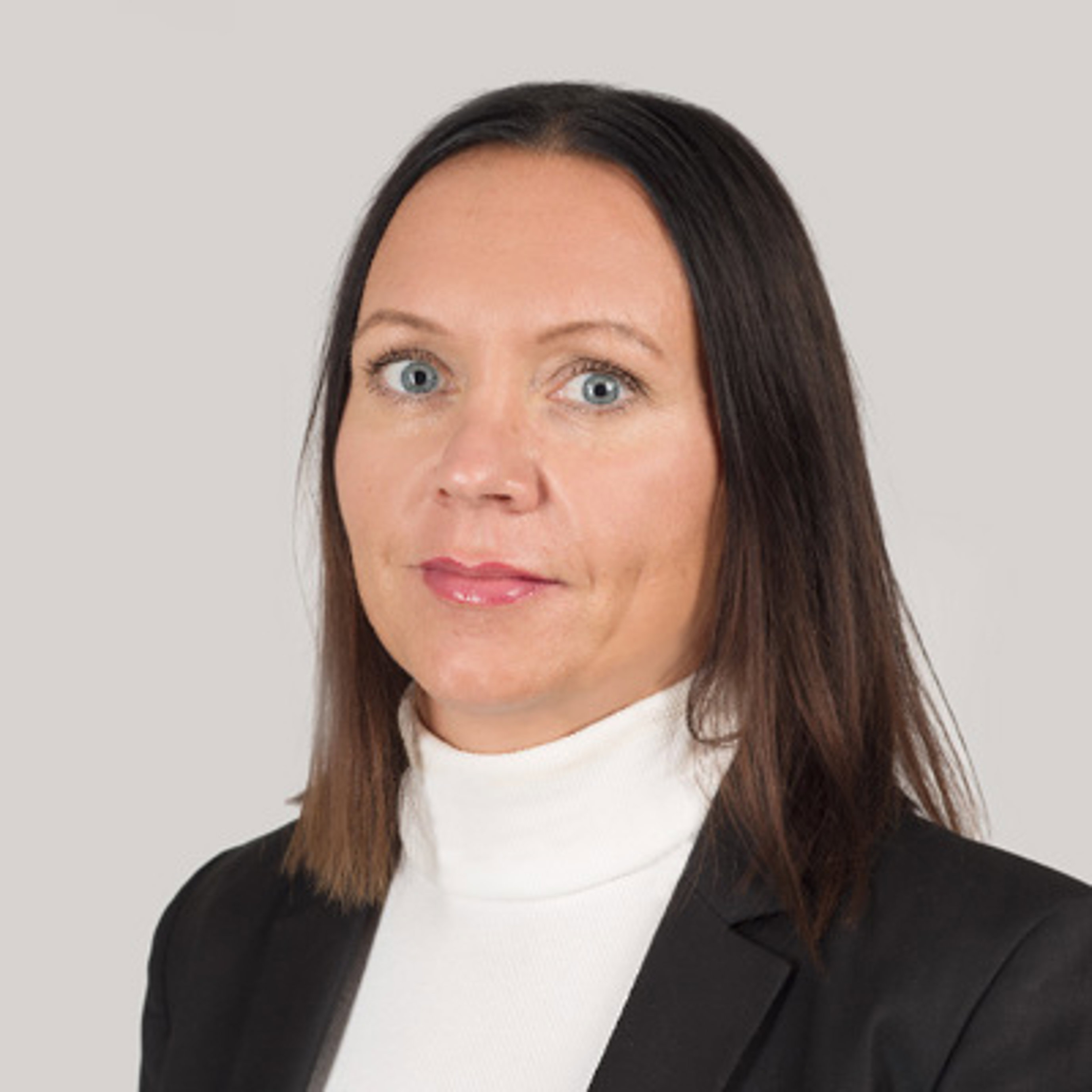 Profesjonell kvinne med langt mørkt hår iført en hvit høyhalset genser og svart blazer, ser direkte på kameraet mot en ensfarget grå bakgrunn