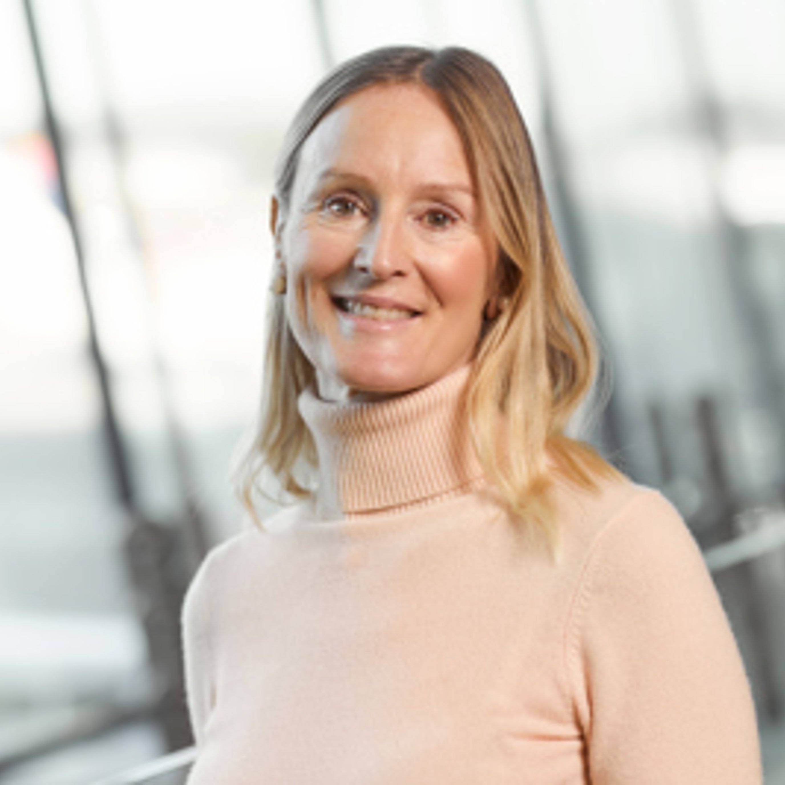 Smilende kvinne i en lys rosa høyhalset genser som står innendørs med en uskarp bakgrunn