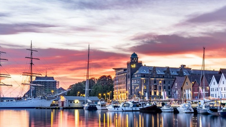 Vakker utsikt over en havn med seilbåter og historiske bygninger ved solnedgang, med livlige himmelfarger og refleksjoner på vannet