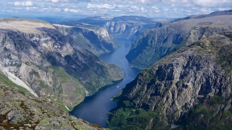 Majestetisk utsikt over en dyp blå fjord omgitt av ruvende steinete klipper og frodige grønne landskap under en klar blå himmel med spredte skyer i Norge