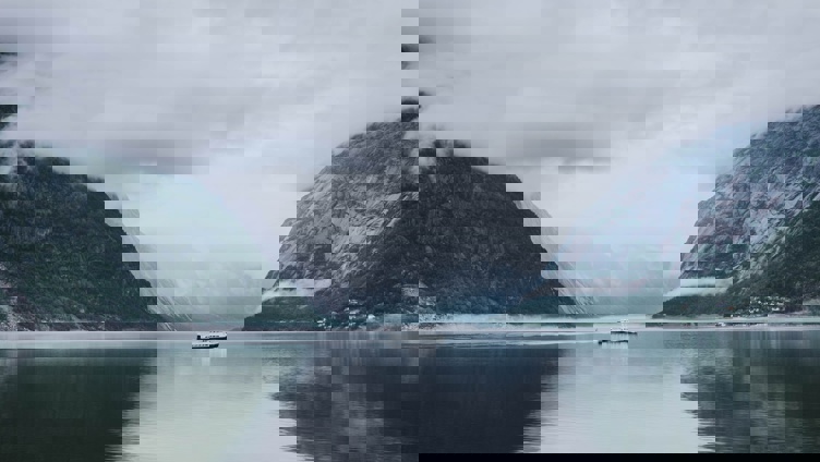 Et rolig landskap med en båt på en stille fjord omgitt av tåkete fjell, under en skyet himmel