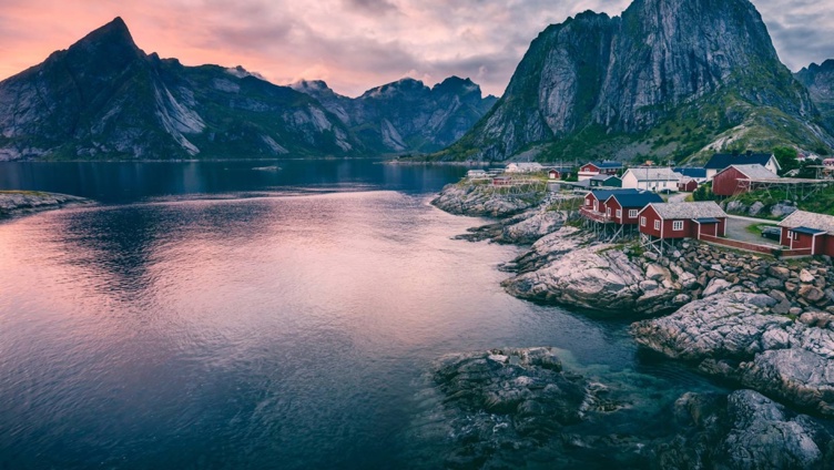 Fjordlandskap i Lofoten med røde rorbuer ved havet og majestetiske fjell i bakgrunnen under en rosa solnedgang.