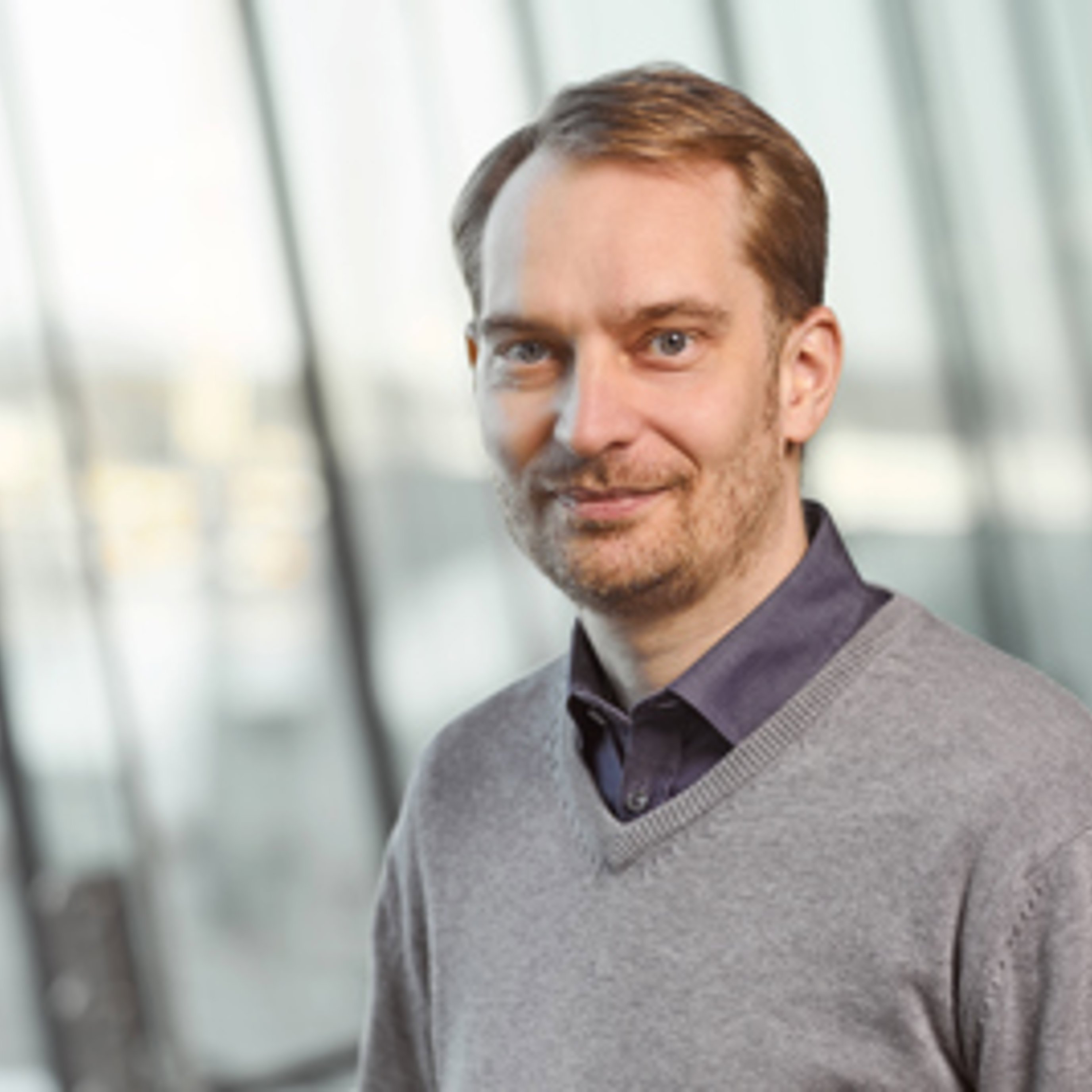 En smilende mann med skjorte og genser står foran en glassvegg.