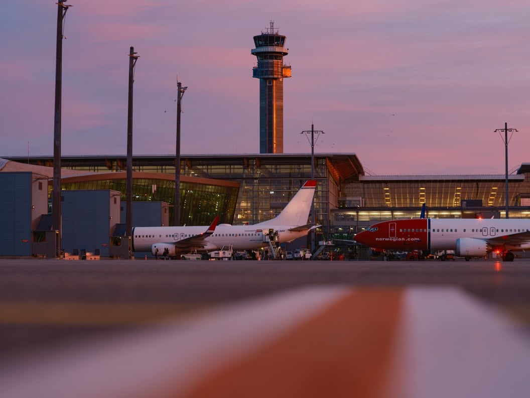 Flyplassomgivelser ved solnedgang med fly som står parkert foran en moderne terminalbygning og et kontrolltårn i bakgrunnen.