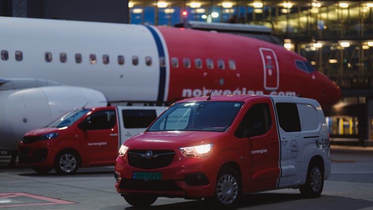 Røde og hvite servicebiler parkert nær et Norwegian Air Shuttle fly ved en flyplassterminal i kveldstimene
