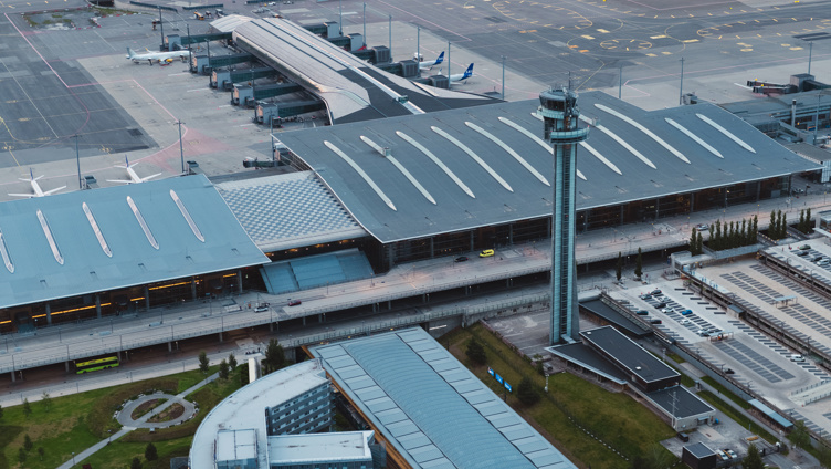 Flyplass med et stort passasjerfly parkert ved terminalen, byggkran i forgrunnen, og søknadsskjema med penn på bordet, symboliserer muligheter innen luftfartsindustrien.