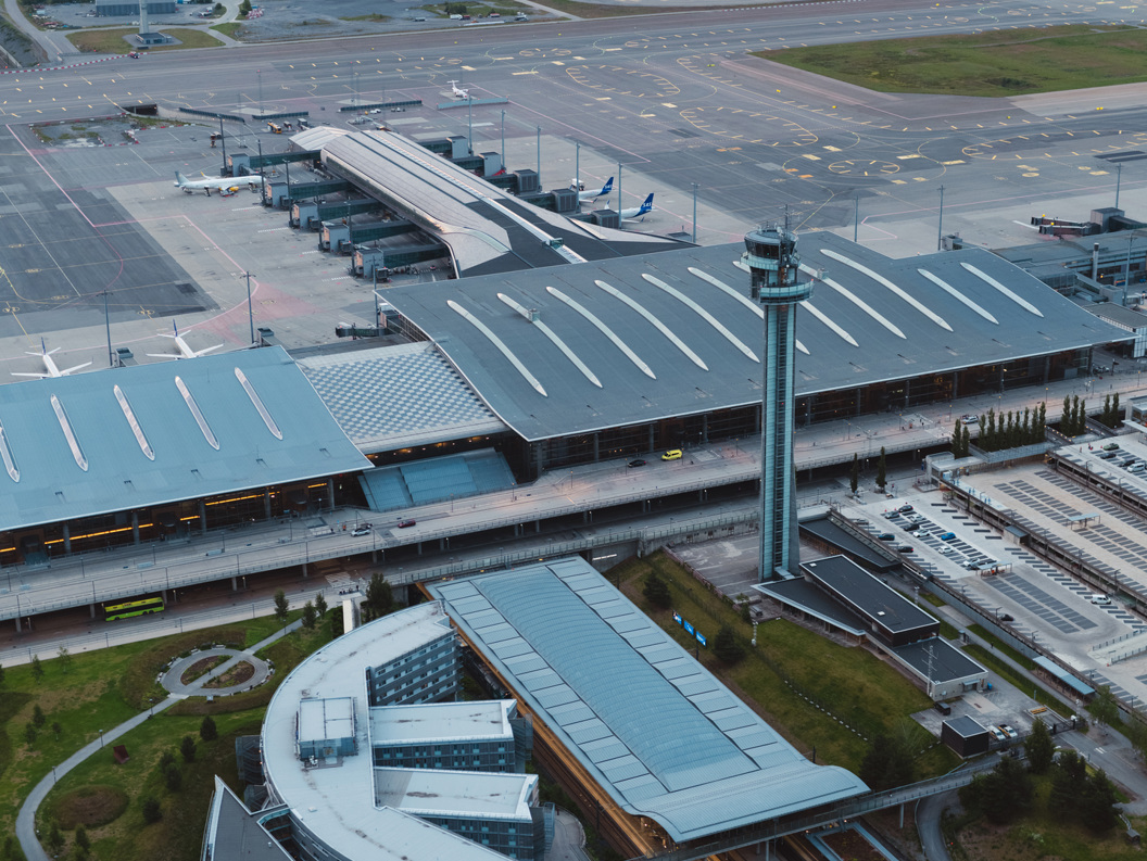 Flyfoto av Oslo lufthavn Gardermoen, med terminalbygg, tårn og rullebaner, omgitt av skog og fjell